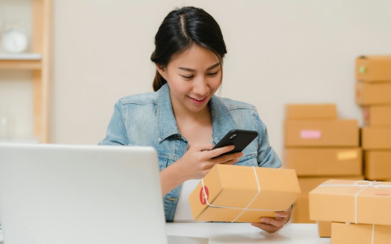 Vendedora online revisando un pedido con el móvil junto a cajas de envío