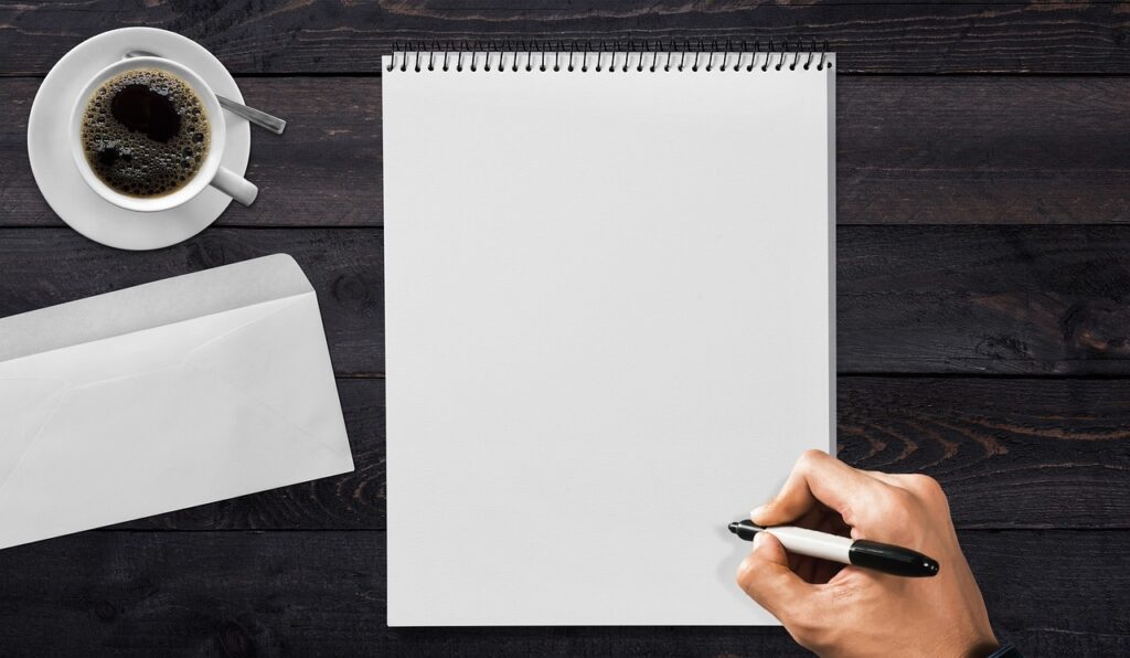Una mano empieza a escribir con un rotulador en un bloc en blanco, junto a un sobre y una taza de café sobre una mesa de madera oscura