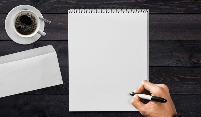 Una mano empieza a escribir con un rotulador en un bloc en blanco, junto a un sobre y una taza de café sobre una mesa de madera oscura
